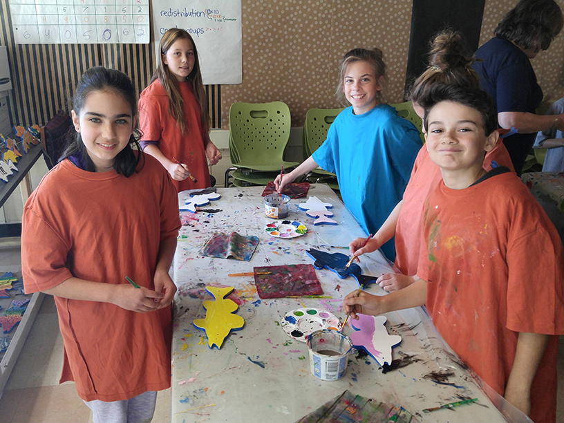 A group of children smile at the camera at a table where they are decorating their wooden fish.