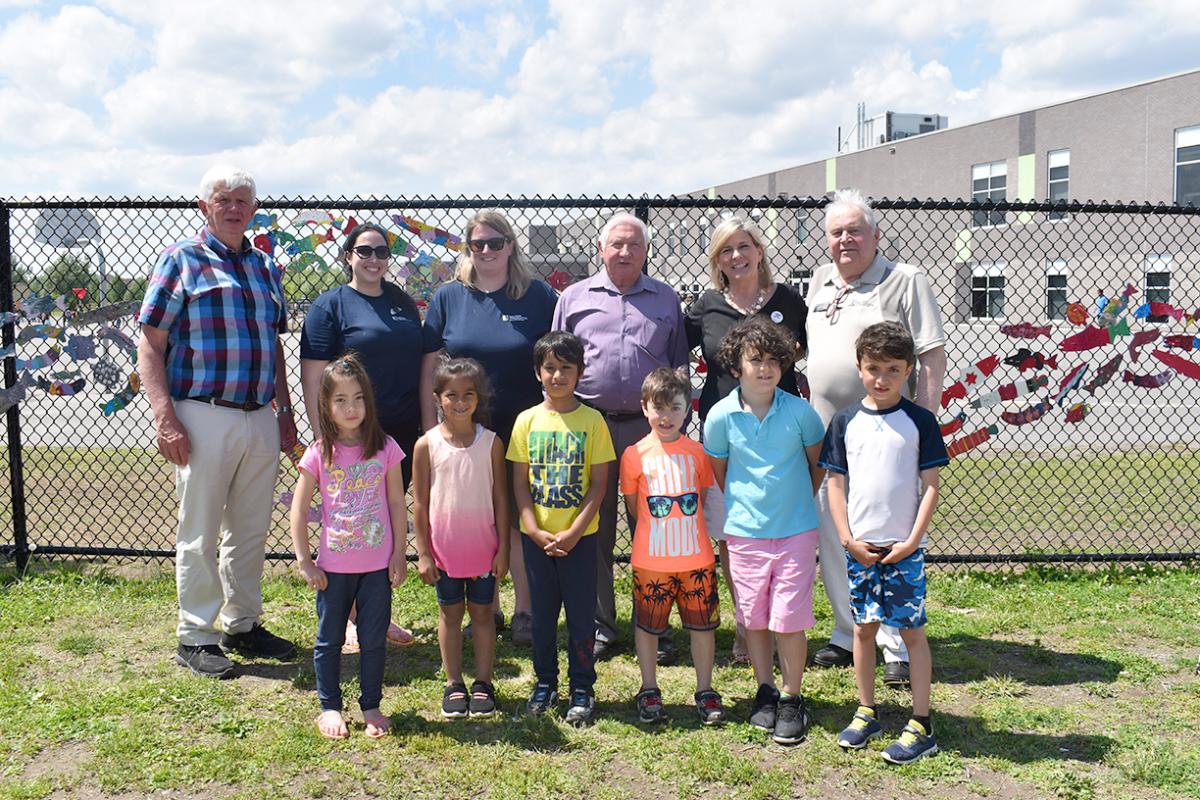 A group of children, SNC employees, teachers, and SNC board members pose in front of a fence with a completed Stream of Dream display