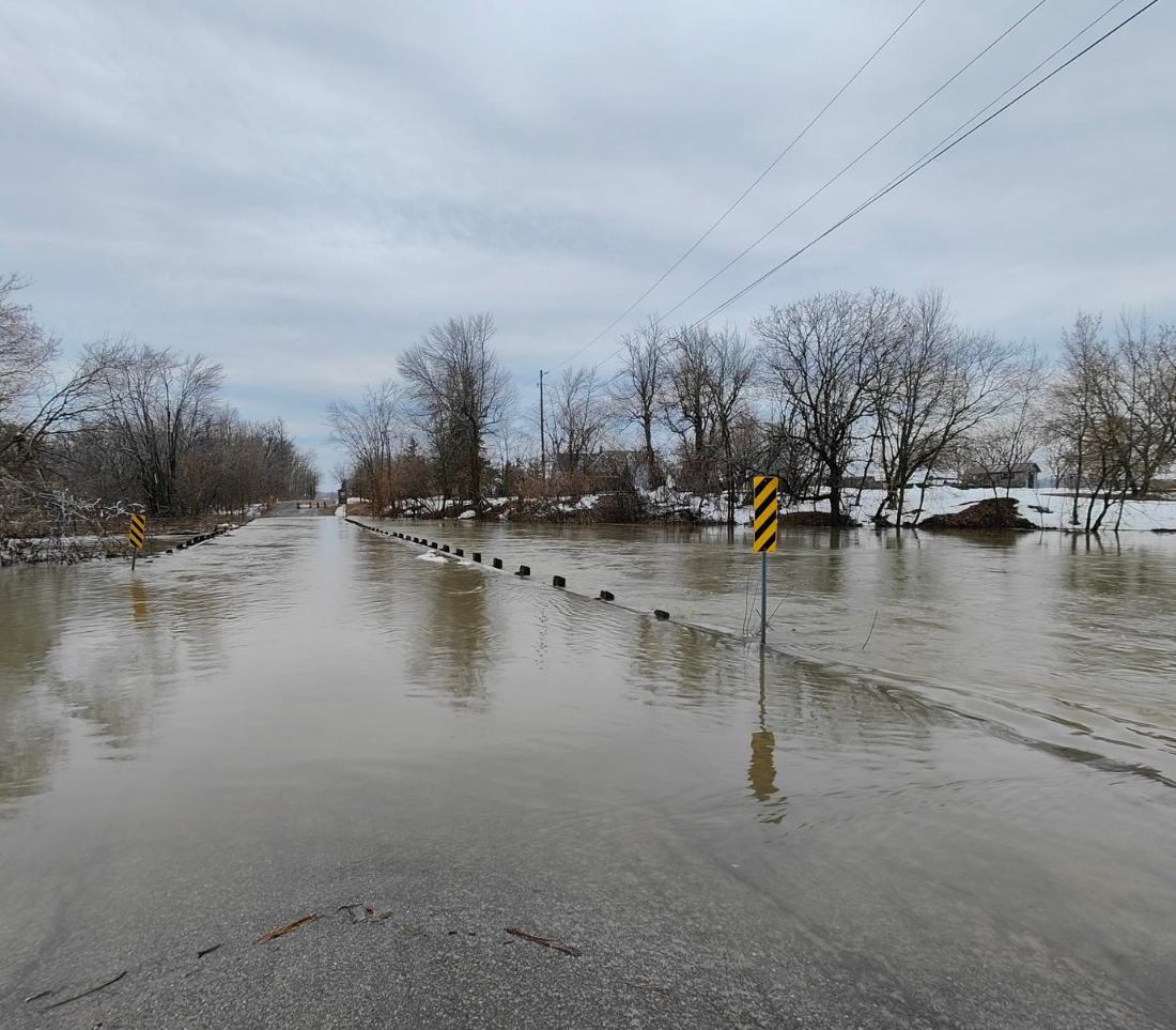 Une photographie prise depuis le bord d'une route asphalt&eacute;e enti&egrave;rement submerg&eacute;e par les eaux troubles de la rivi&egrave;re South Castor en crue. Des panneaux de signalisation ray&eacute;s jaune et noir se dressent dans l'eau pour marquer le trac&eacute; de la route. L'arri&egrave;re-plan montre des arbres d&eacute;nud&eacute;s, des plaques de neige fondante sur les berges et un ciel gris et couvert.